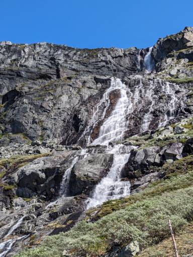 En vakker alpefoss som renner nedover mange store steiner i Norge, dagen er varm og himmelen er blå uten skyer.