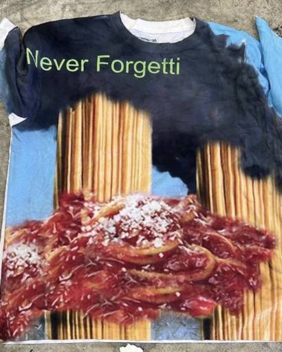 A shirt that has two “towers” of uncooked spaghetti with smoke coming out of them, and some cooked spaghetti in marinara sauce with parmesan on top layered onto the towers. the whole scene is placed on a blue sky background and on the black smoke in the upper left, there is a caption saying “Never Forgetti”.

It's the twin towers made out of spaghetti.